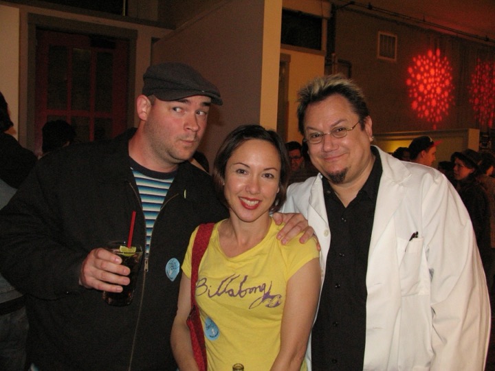 Three friends pose at a crowded party: a man in a flat cap holding a drink, a smiling woman in a yellow T-shirt, and a man with bleached hair in a white lab coat, red lights on the wall behind.