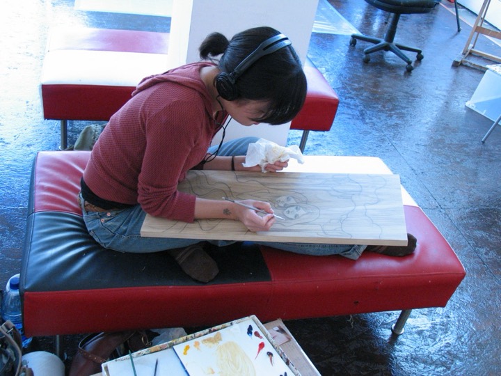 An artist in headphones sits on a red bench drawing two faces on a wood panel.