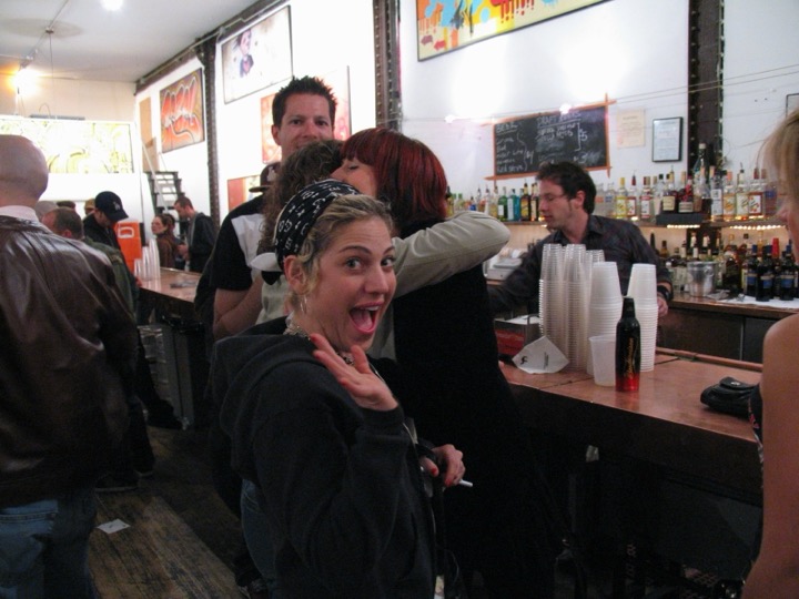 At a gallery bar, a woman in a bandana waves at the camera with her mouth open in mock surprise while friends hug behind her.
