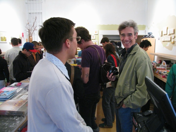 A busy shop-and-gallery opening: people browse books and zines while a smiling man holding a camera looks toward the lens.