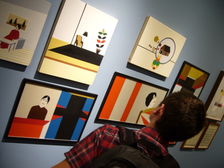 A visitor looks at flat, brightly colored paintings of rooms and women hung on a blue wall.