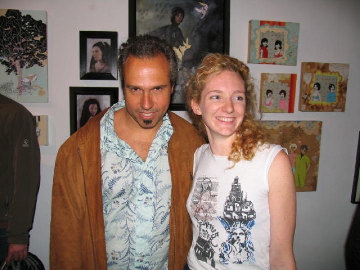 A man in a suede jacket and a smiling woman with curly red hair pose in front of small paintings.