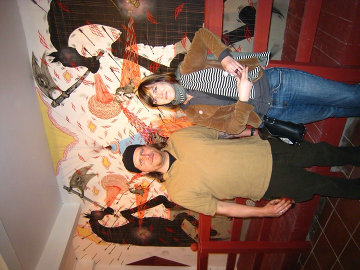 Two people stand in front of a huge mural of black horses, axes and arrows.