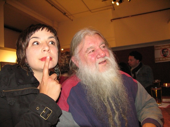 A woman holds a finger to her lips beside a man with a long white beard.