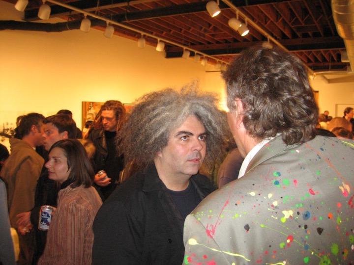 Two men talk face to face in a packed gallery under track lights; one has a big cloud of gray curly hair, the other wears a paint-splattered jacket.