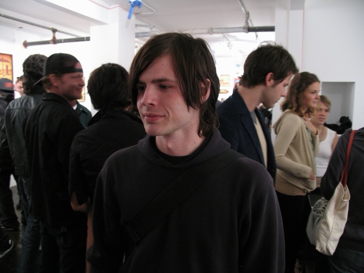 A young man with long bangs stands in a crowded white gallery.