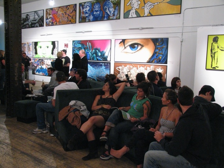 People lounge on long sofas at an opening, under a wall of big graffiti-style paintings.