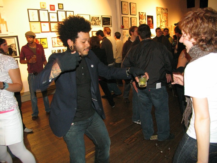 A man in a blazer dances with a drink in his hand in a packed gallery hung salon-style.