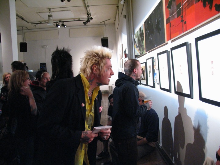 A man with spiked blond hair and neck tattoos studies framed work on a gallery wall.