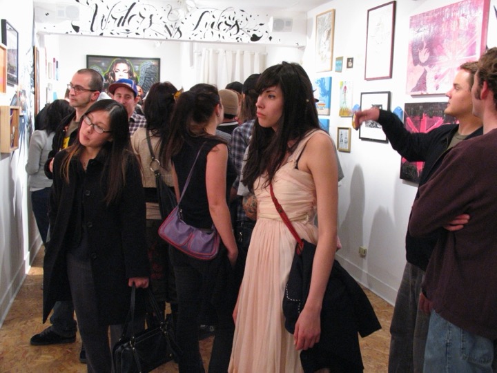 A crowd fills a narrow white gallery hung salon-style, with hand-lettered script painted above the doorway.