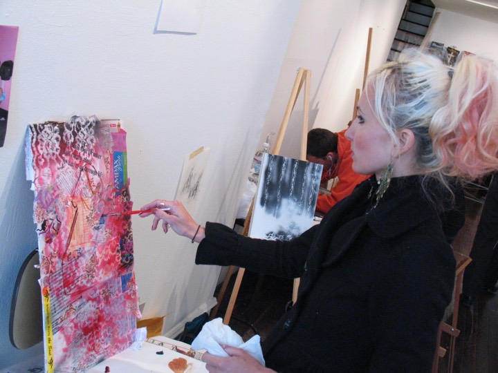 A woman with pink and blonde hair paints a pink collage on an easel.