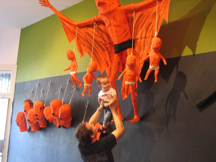 A man lifts a baby up toward a giant orange winged figure and hanging orange figures on a gallery wall.