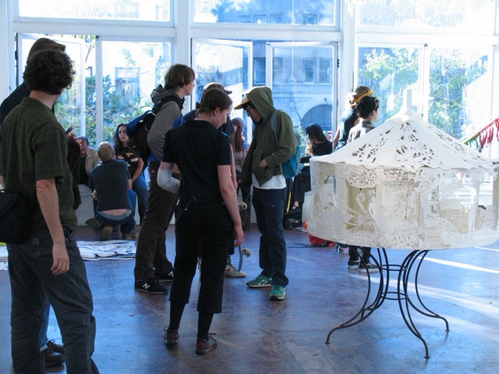 Visitors stand in a sunny storefront gallery beside an intricate white cut-paper carousel on a wrought-iron stand.