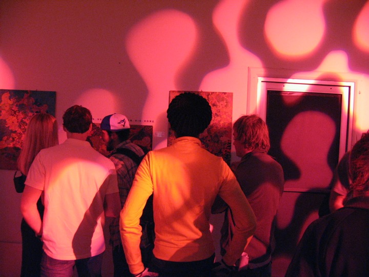 People look at paintings in a room washed with pink and orange light patterns.