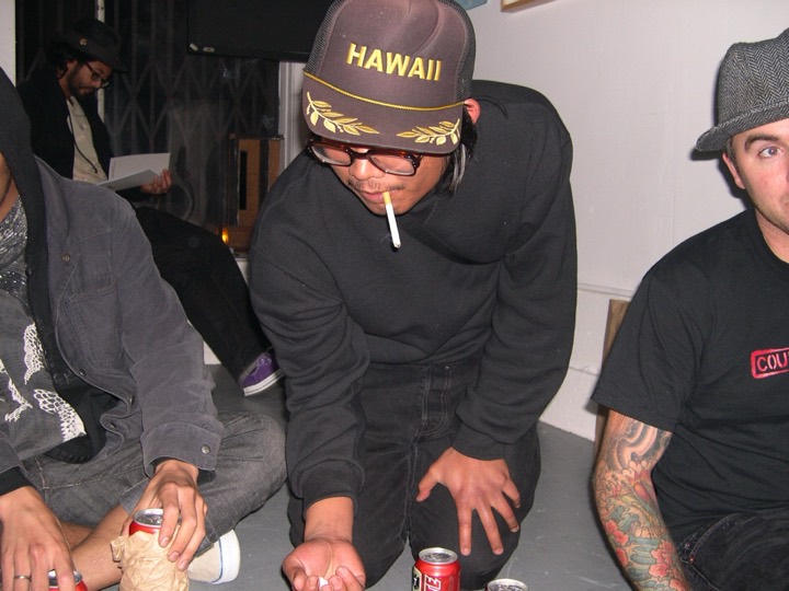 Men crouched on a gallery floor among beer cans, one in a Hawaii trucker cap with a cigarette, rolling dice.