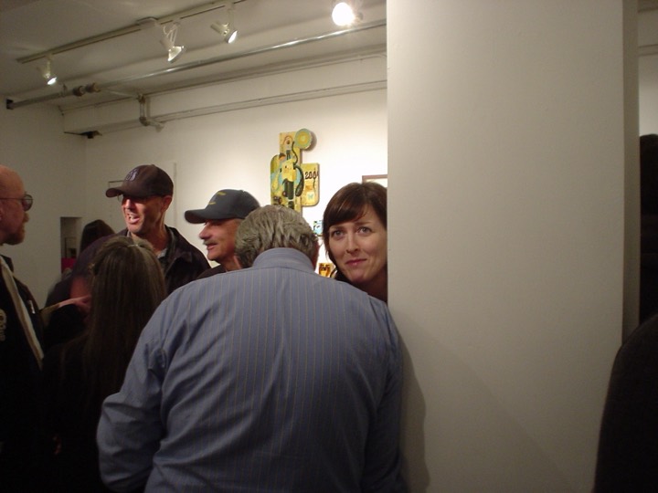 A crowded gallery opening under track lights; one person peeks around a white wall at the camera.