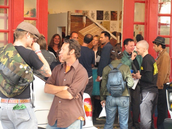 An opening spills out of a gallery with red doors onto the sidewalk, people talking in small groups.