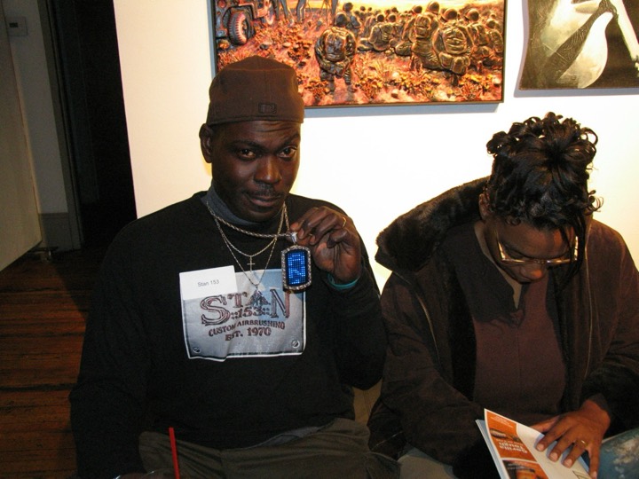 A man in a Stan 153 Custom Airbrushing shirt holds up a glowing blue pendant.