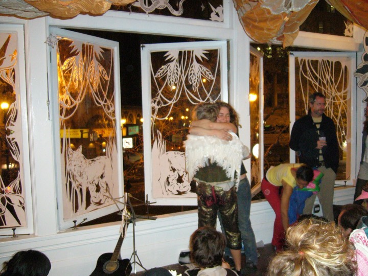 Two people hug in front of windows hung with white cut-paper wolves and plants, one wearing feather wings.