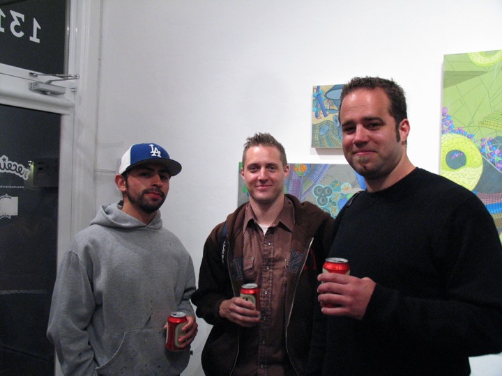 Three men holding beer cans pose by a gallery door, colorful geometric paintings on the wall behind them.